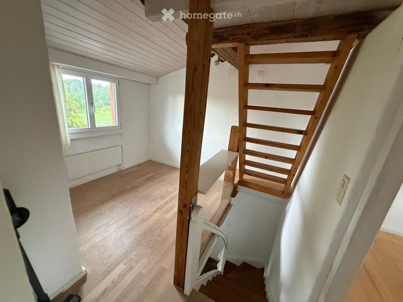 Empty room with wooden flooring, white walls, and a window showing greenery outside. A staircase leads upwards with wooden railings.