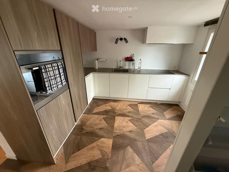 Modern kitchen with white cabinets, wooden herringbone floor, stainless steel oven, and sink area.