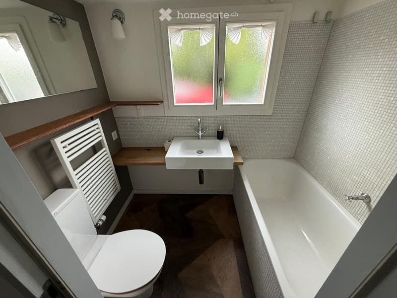 Modern bathroom with a white toilet, bathtub, sink, and radiator. The room features grey tiled walls, wooden flooring, and a window with frosted glass.