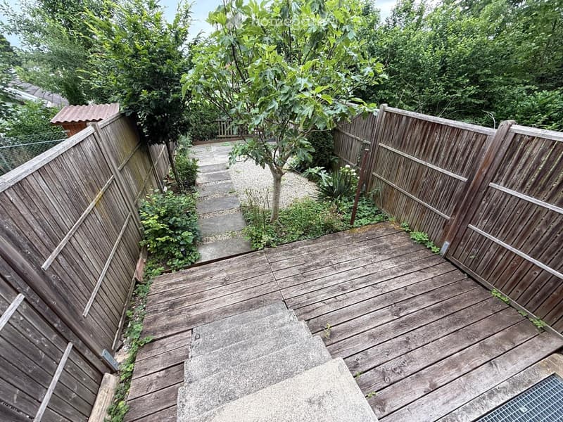 Wooden deck with surrounding wooden fences, small garden area with plants and a tree, stone pathway leading to a raised section.
