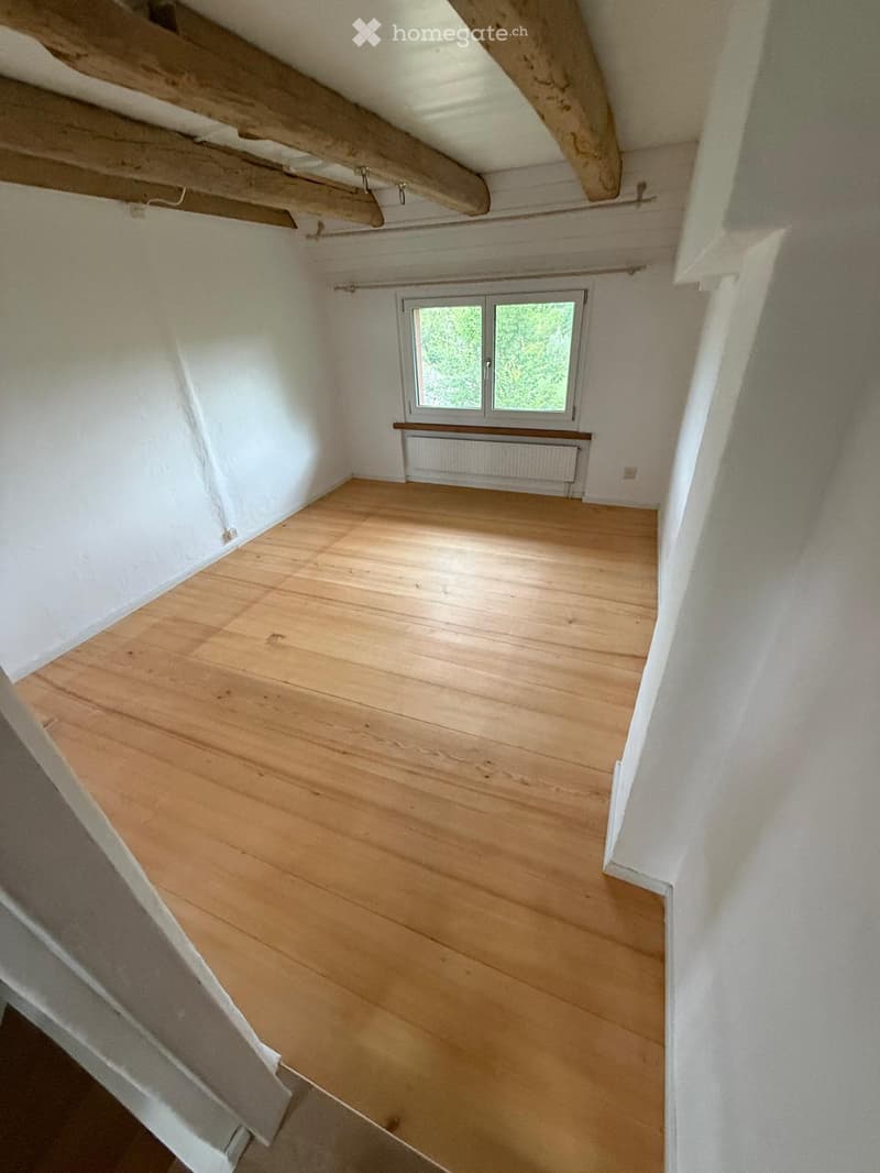 Empty attic room with wooden flooring, white walls, exposed wooden beams, and a window with a radiator below it.