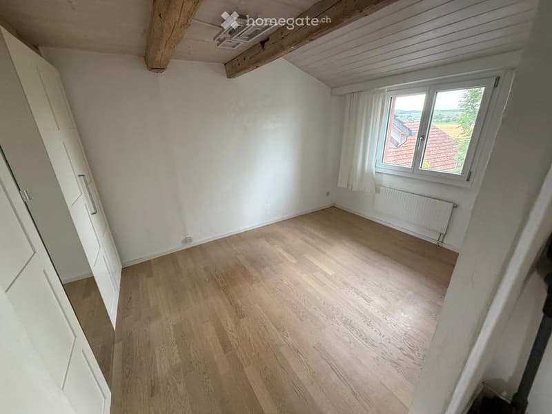 Empty attic room with wooden flooring, white walls, and a large window offering a view of the countryside.