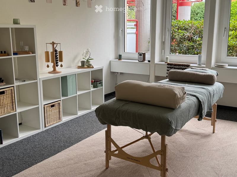 A well-lit room with a massage table, shelves, decorative items, and a view of greenery outside.