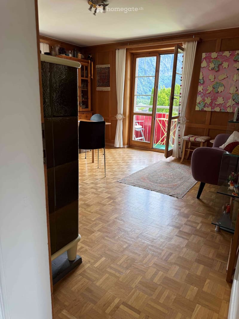 A spacious living room with wooden herringbone flooring, a large window offering a scenic mountain view, a sofa, a chair, a small table, a stool, a cabinet, a mirror, and a colorful painting on the wall.