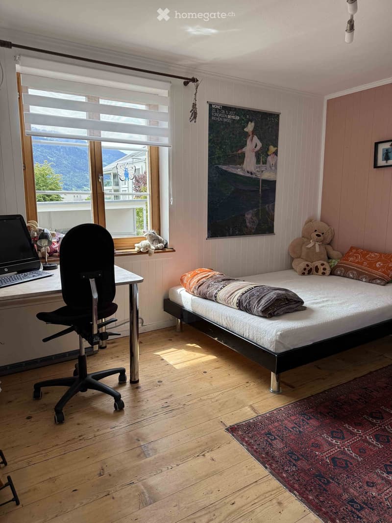 Cozy bedroom with a single bed, desk, chair, and a view of the mountains through large windows. The room features wooden flooring, a colorful rug, and decorative pillows on the bed.