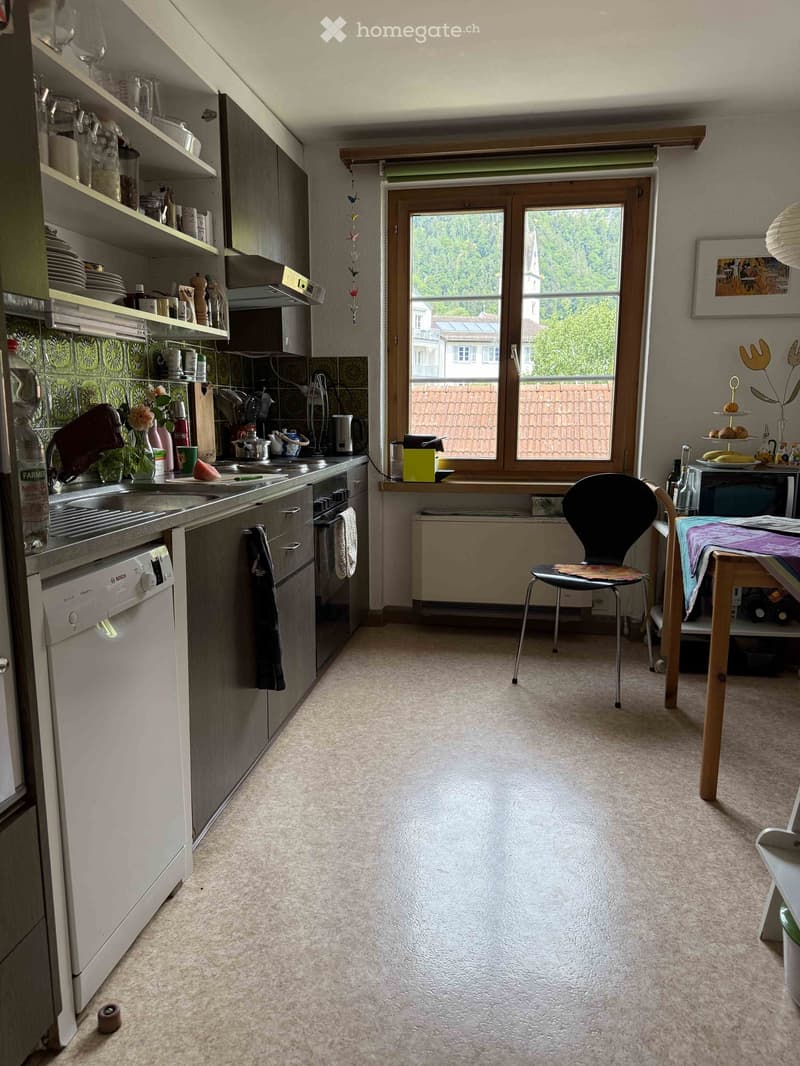 Modern kitchen with grey cabinets, white appliances, and a large window overlooking a garden. Includes a dishwasher, oven, sink, fridge, and various kitchen items on the counter and shelves.