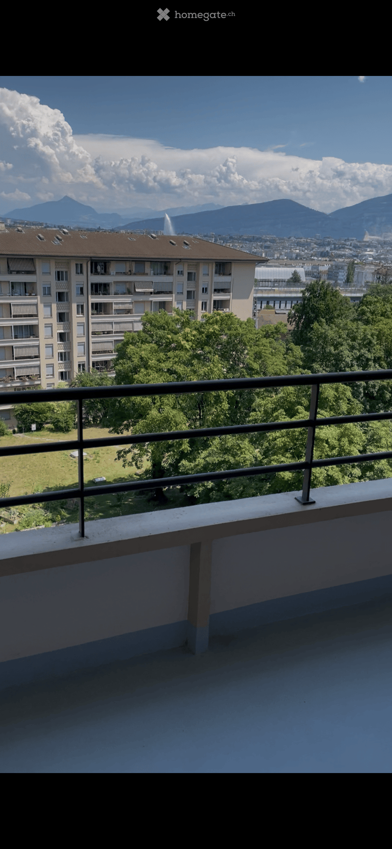 Balcony with metal railing, overlooking a park and cityscape with mountains in the background.
