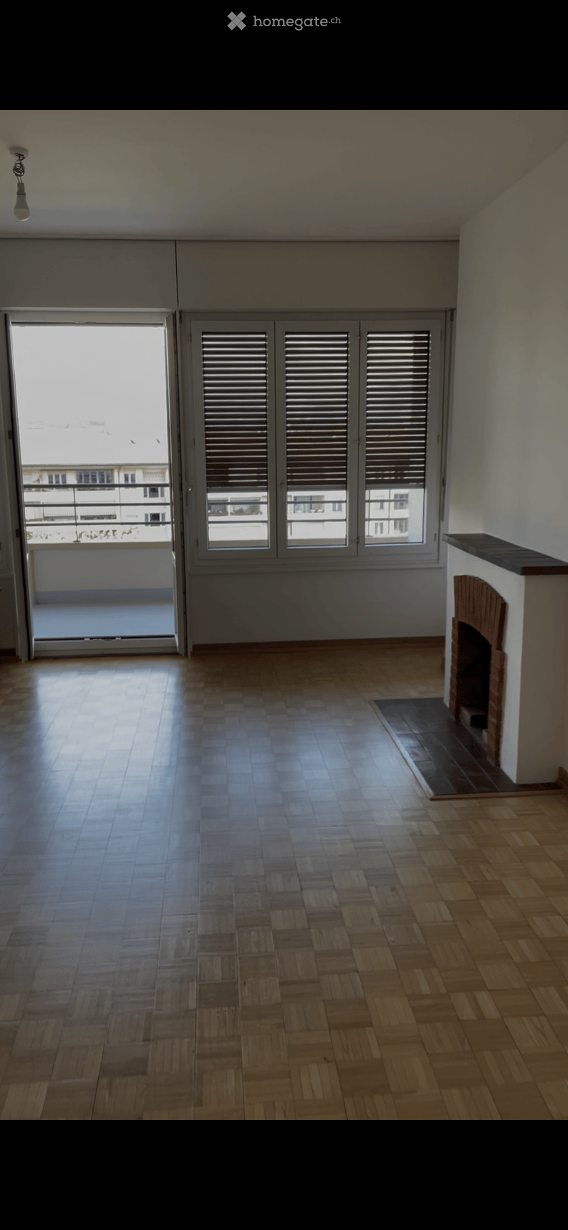 Empty room with wooden flooring, fireplace, large windows with shutters, and a balcony view.