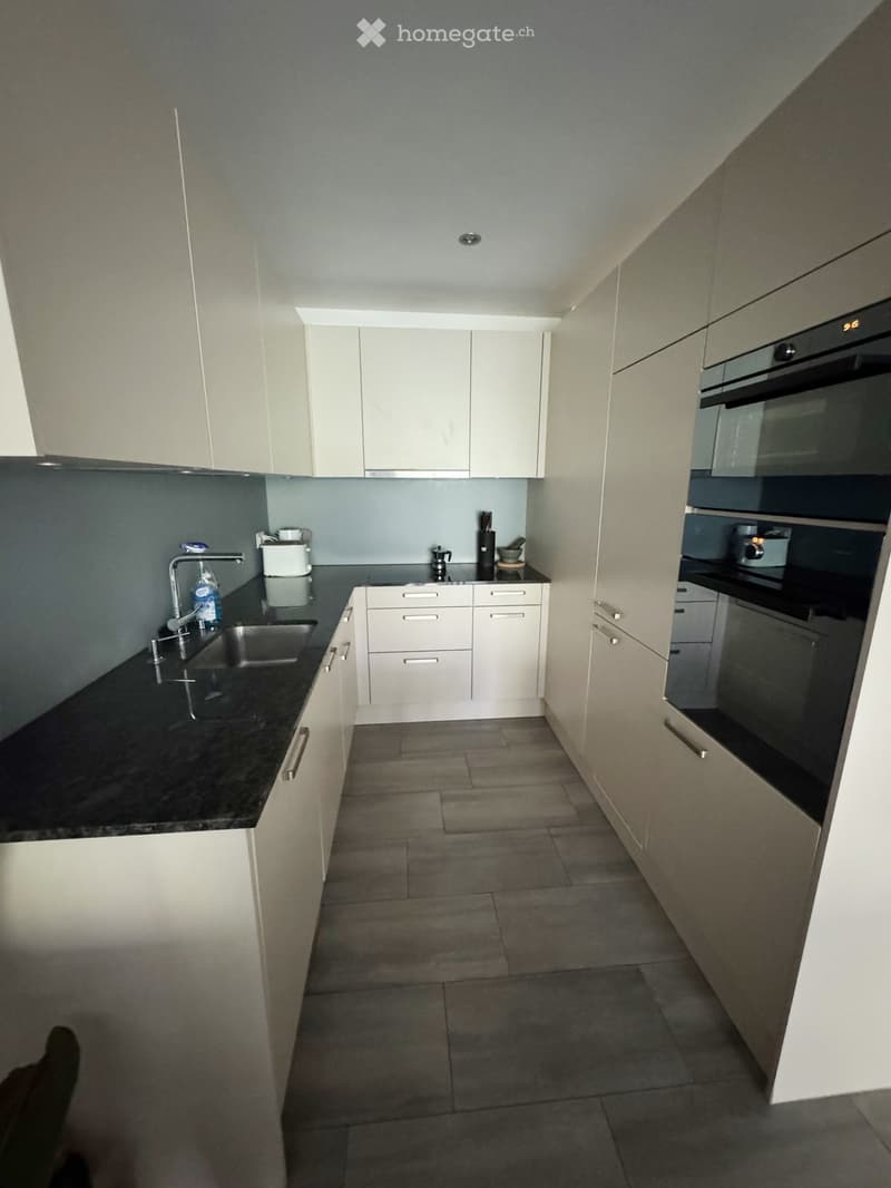 Modern kitchen with white cabinets, black countertops, and stainless steel appliances including a sink, faucet, and oven. The floor is tiled, and the space is well-lit with natural light from the left side.