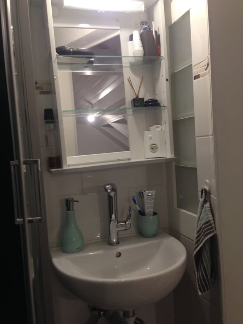A modern bathroom with a white sink, chrome faucet, and a mirrored cabinet with glass shelves holding toiletries. A small green soap dispenser and toothbrush holder are on the sink counter, and a striped towel hangs on a rack.