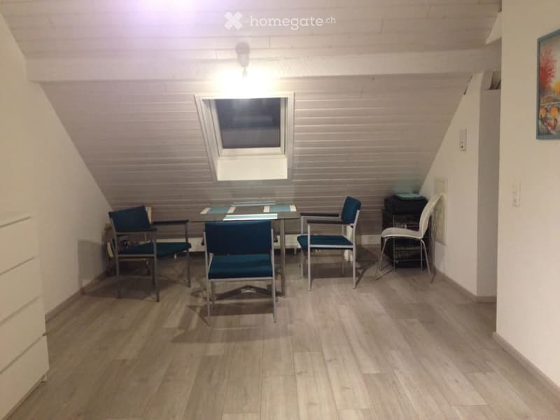 A small, modern dining area with a glass table, four chairs, and a stool, featuring wooden flooring and white walls.
