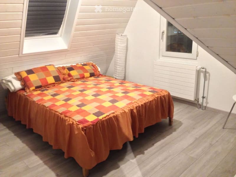 Cozy attic bedroom with wooden flooring, slanted white paneled ceiling, and large windows. Features a single bed with a colorful patchwork quilt and matching pillows, a radiator, and a small stool.
