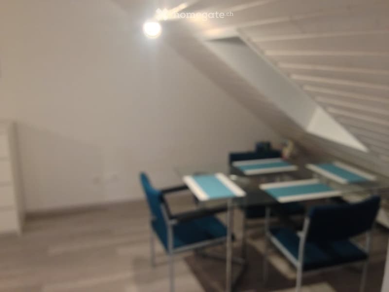 A small, modern room with a glass table and blue chairs, likely a dining or study area, featuring white walls and a slanted ceiling.