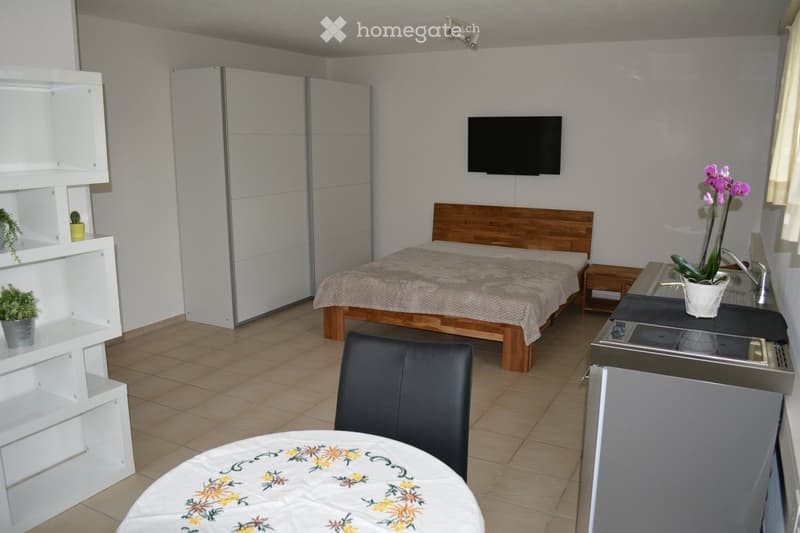 A compact, furnished room with a bed, a small kitchen area, a TV, a chair, and a table with a floral tablecloth.