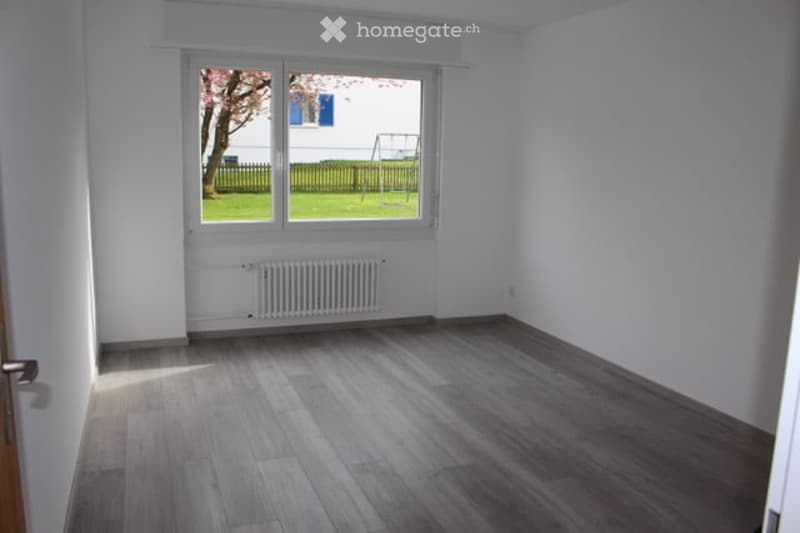 Empty room with wooden flooring, white walls, large window with view of a green yard, radiator below the window, and a door on the left side.