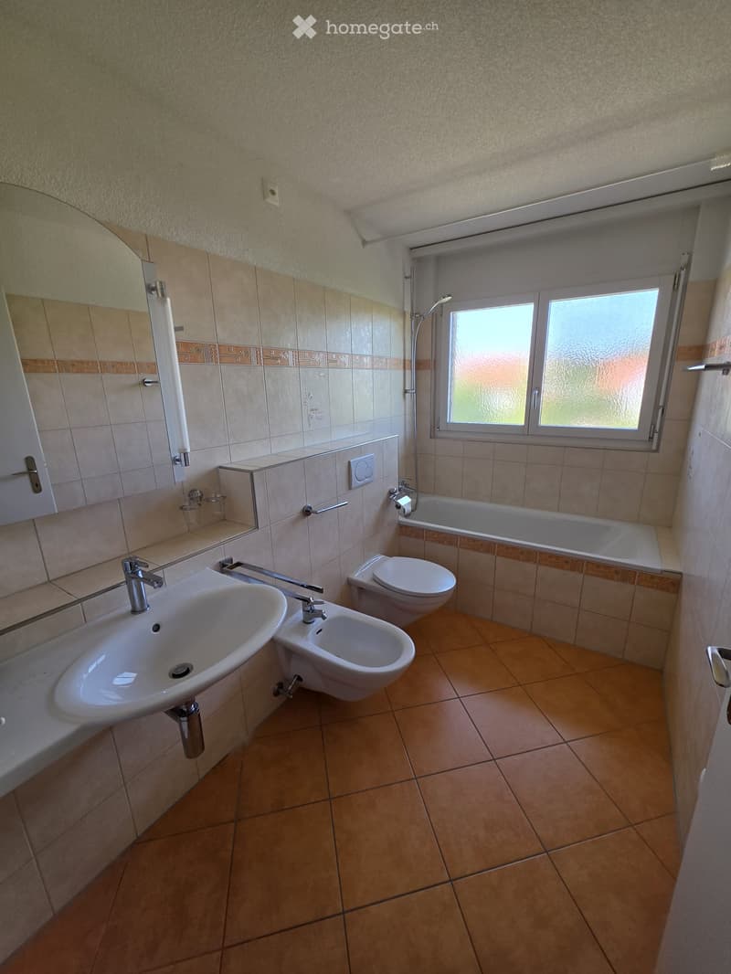 Modern bathroom with beige tiles, a sink, toilet, bidet, bathtub, and a large window providing natural light.