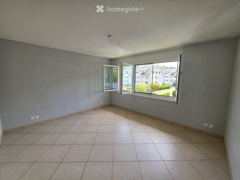 Empty room with tiled floor, grey walls, and large windows offering a view of the outside area.