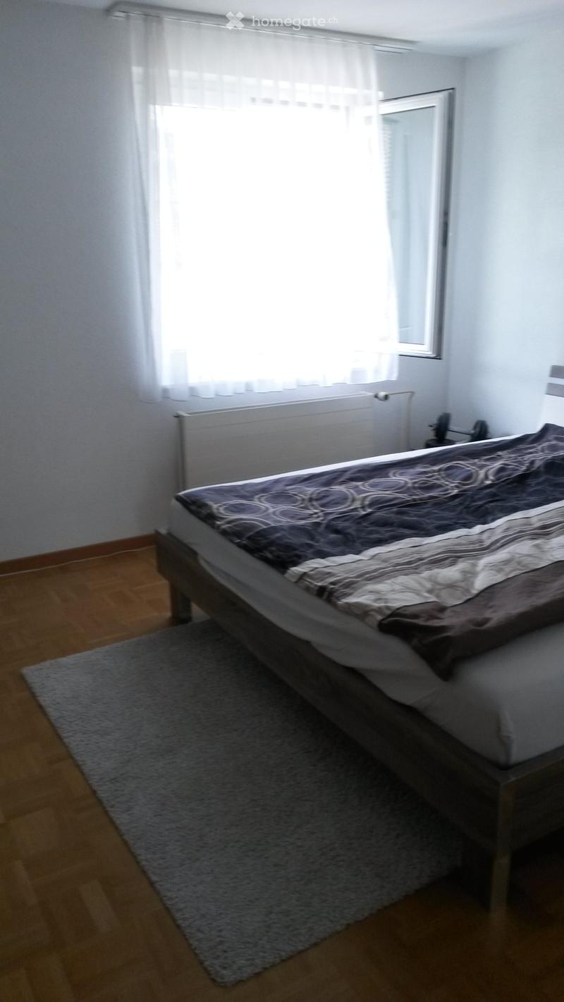Bedroom with wooden flooring, a bed with dark bedding, a gray rug, and a window with white curtains.