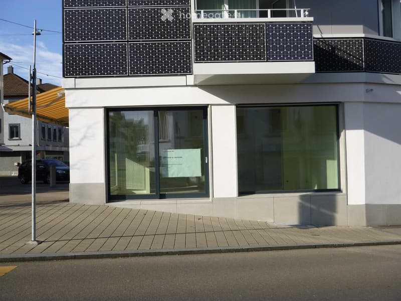 Modern two-story building with large glass windows and a balcony on the upper floor. The ground floor has a storefront with glass doors and a sign. The building features a clean, contemporary design with white walls and grey accents.