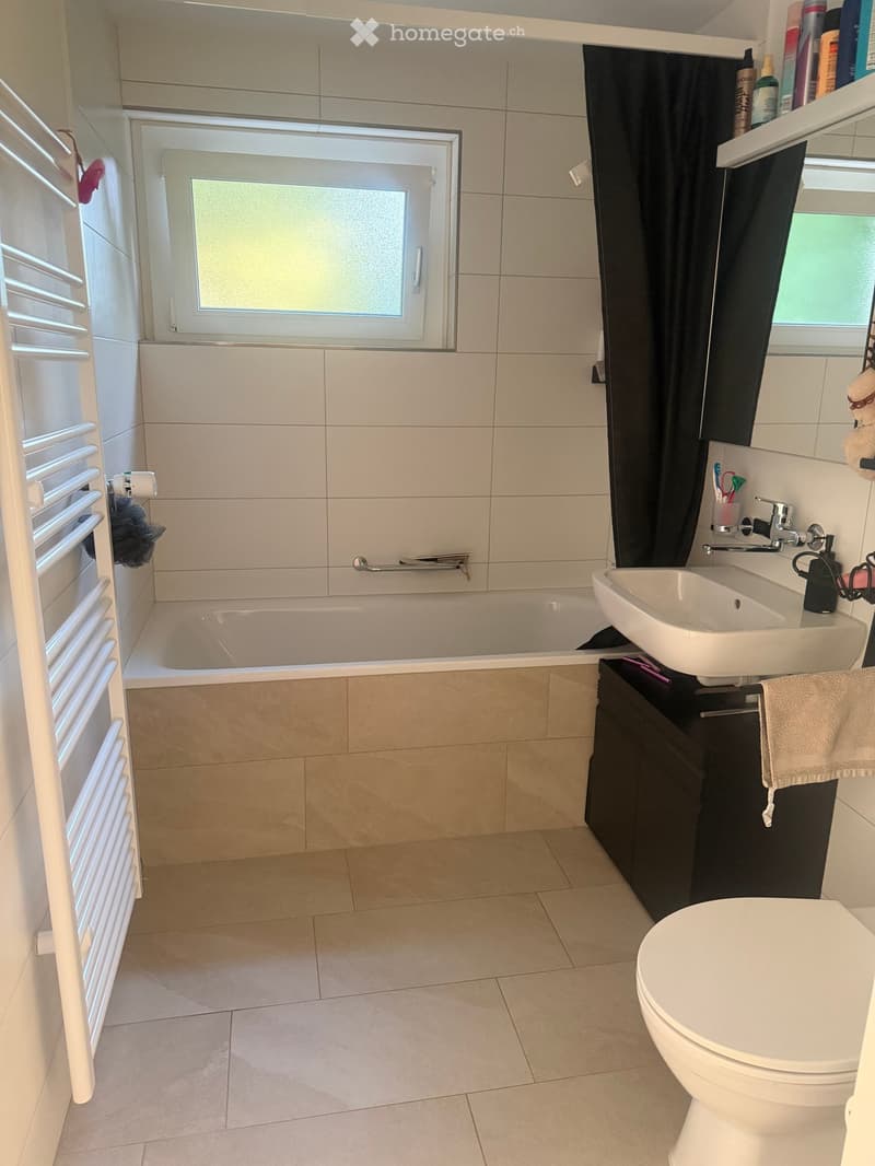 Modern bathroom with a bathtub, sink, toilet, and a radiator. Features tiled walls and floor, a window with frosted glass, and a black shower curtain.