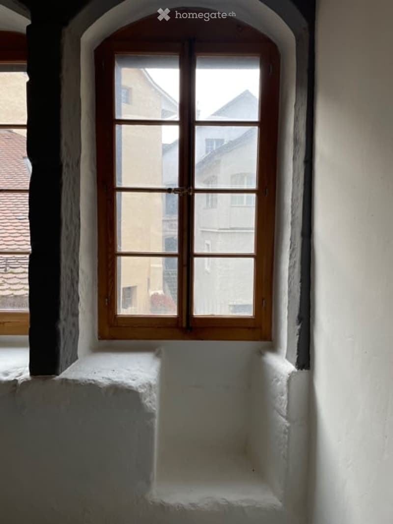 An empty room with a single window and a small stone bench beneath it. The walls are white and the window frame is wooden.