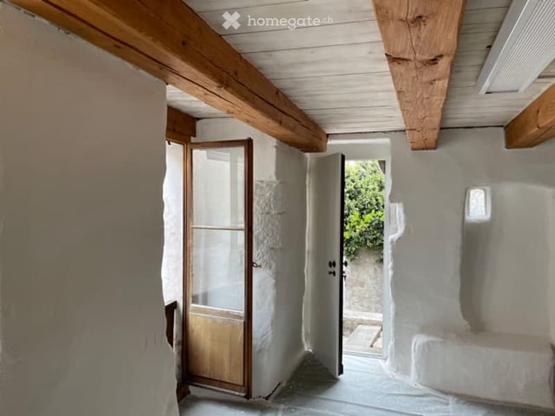Empty room with white plaster walls, wooden ceiling beams, and a glass door leading outside.