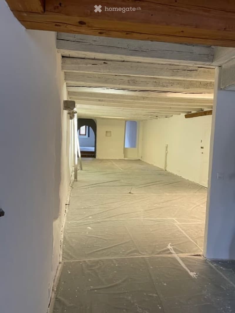 Empty room with bare walls and a wooden ceiling, showing a protective plastic sheet on the floor and a partially visible adjacent room.