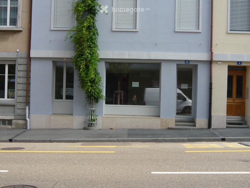 A two-story building with a light gray facade, multiple windows, and a small green plant climbing up the wall. The ground floor has a large window and a door, with a small staircase leading to the entrance.