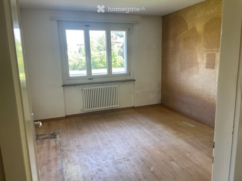 Empty room with wooden flooring, white walls, a window with white frames, and a radiator below the window.