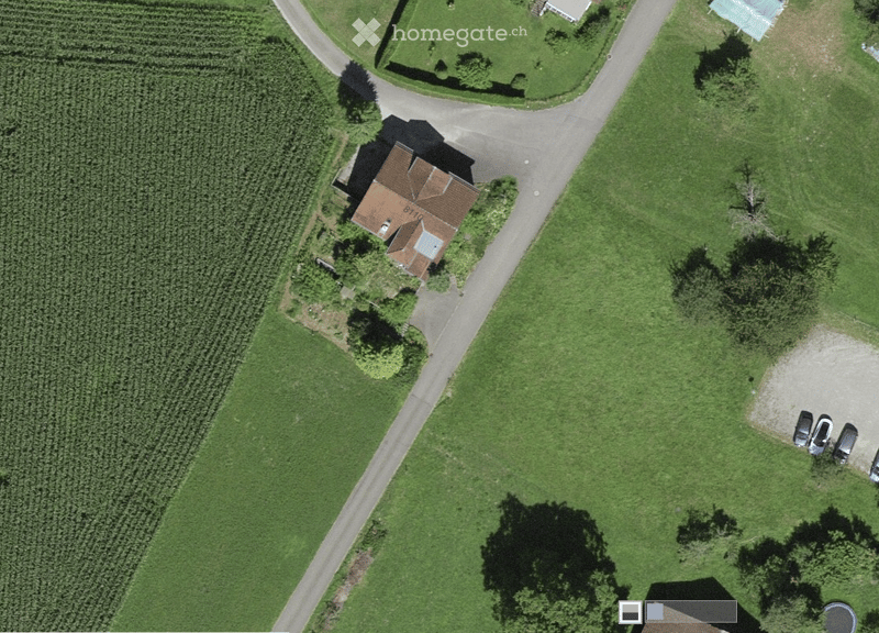 Aerial view of a single-story house with a brown roof, surrounded by green fields and a driveway leading to a parking area with several cars.