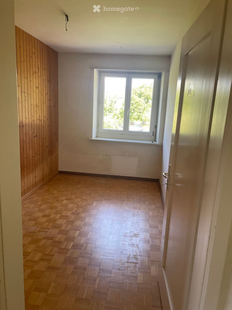 Empty hallway with wooden flooring, white walls, and a large window at the end, allowing natural light to enter.