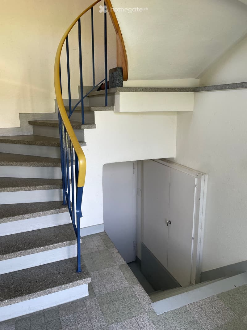Staircase with blue and yellow handrail, gray tiled floor, white walls, and a closed white door leading to another area.