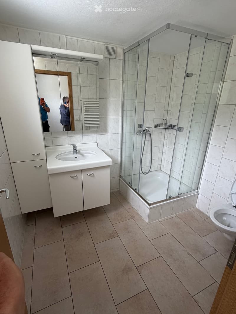 Modern bathroom with a white sink, mirror, glass-enclosed shower, and toilet. The walls are tiled, and the floor is covered with beige tiles.