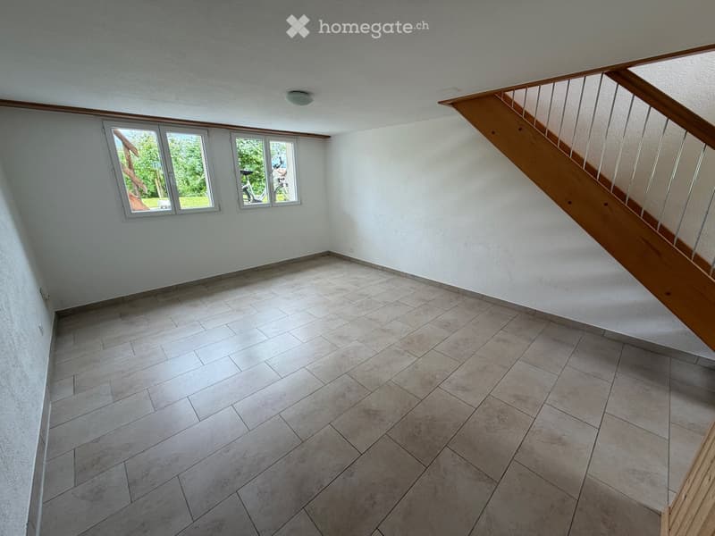Empty room with tiled floor, white walls, and a wooden staircase leading upwards. Two windows on one wall provide natural light.