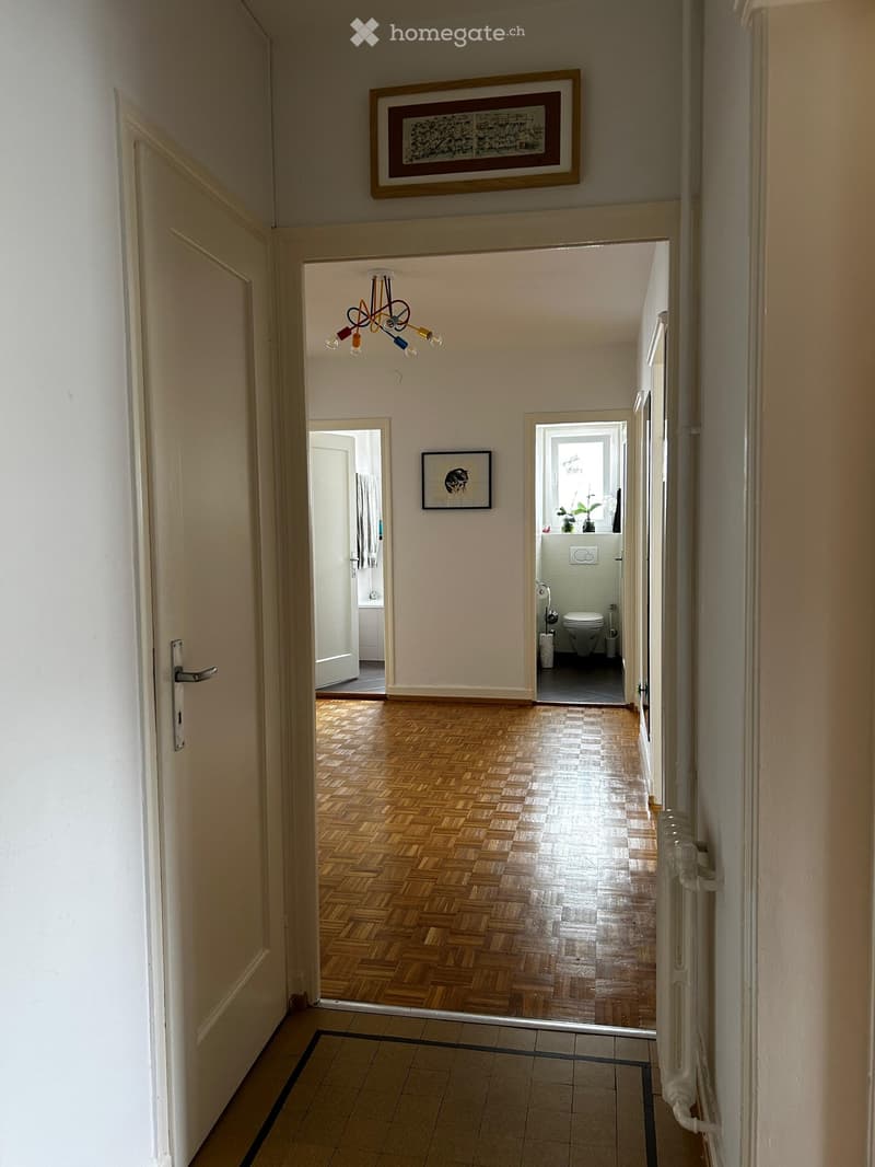 Hallway with wooden flooring, white walls, and a chandelier. Doors lead to a bathroom and another room. Artwork is displayed on the walls.
