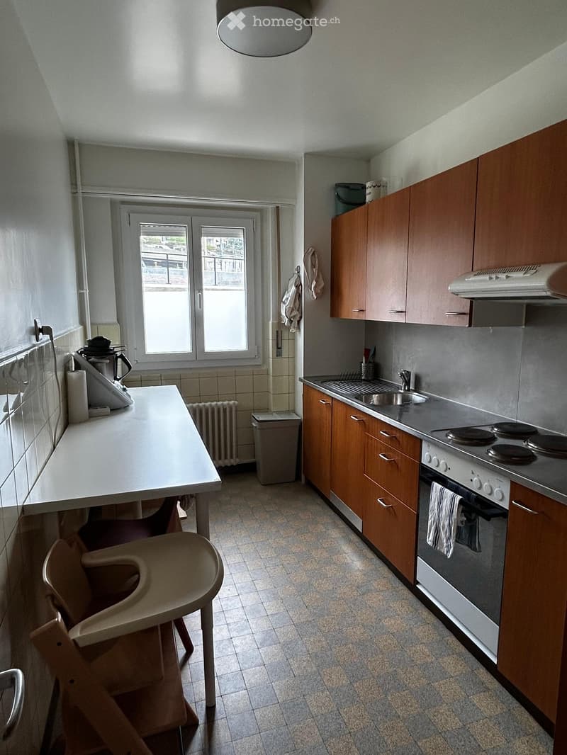 A small kitchen with wooden cabinets, a white countertop, a sink, a stove, and a small table with a chair. The room has tiled floors and walls, with a window providing natural light.