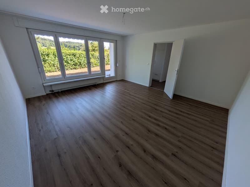 Empty room with wooden flooring, large windows, and an open doorway leading to another space.
