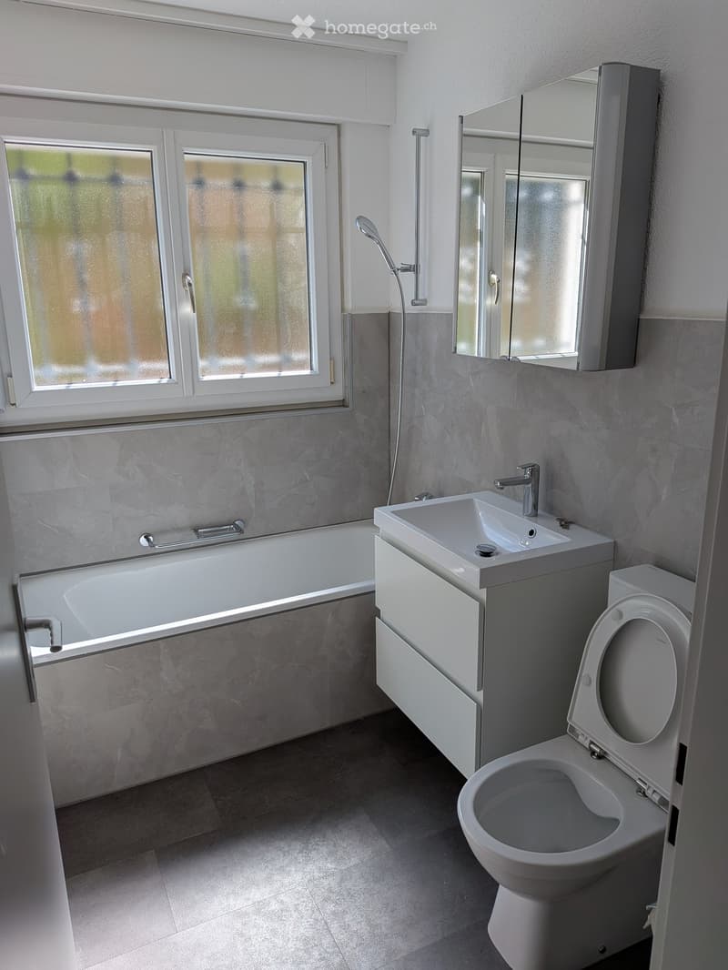 Modern bathroom with a bathtub, sink, toilet, and a large mirror above the sink. The room features grey tiled walls and a window with frosted glass.