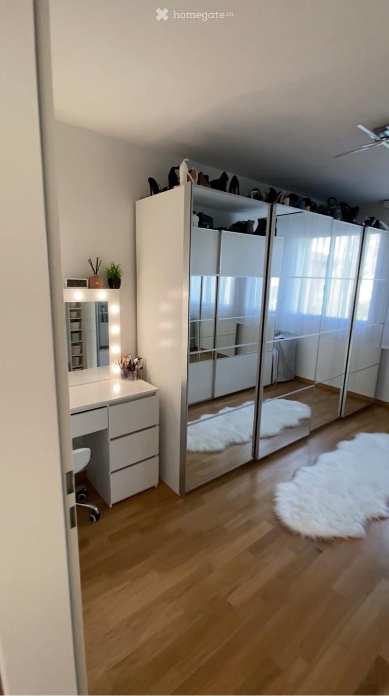 A well-lit, modern walk-in closet with a large mirrored wardrobe, a vanity desk with a mirror, a small plant, and a white fur rug on the wooden floor.