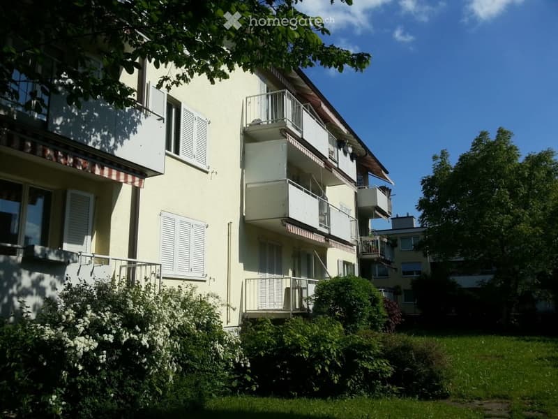 Multi-story residential building with balconies, white shutters, and greenery in front.