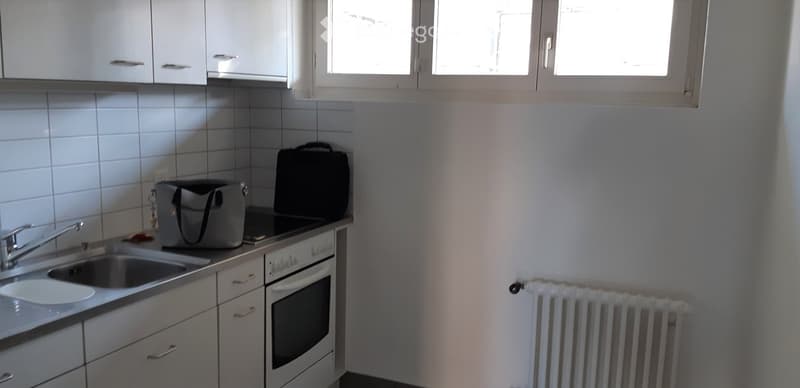Compact kitchen with white cabinets, a sink, an oven, and a radiator. Two bags are placed on the counter.