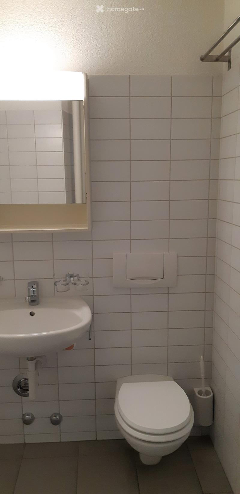 A small bathroom with white tiled walls, a white sink with a chrome faucet, a toilet, and a toilet paper holder. The floor is tiled in a dark color.