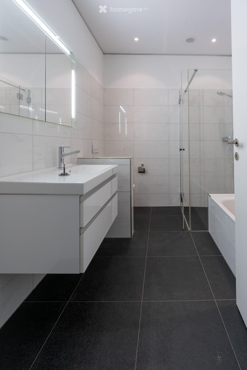 Modern bathroom with white tiles, black floor, large mirror, sink with chrome faucet, and glass-enclosed shower stall.