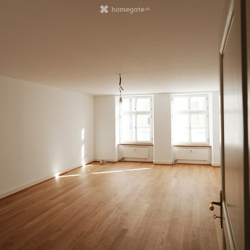 Empty room with light wooden flooring, white walls, and two windows with white frames and blinds. Radiators are visible beneath the windows, and a single light fixture hangs from the ceiling.