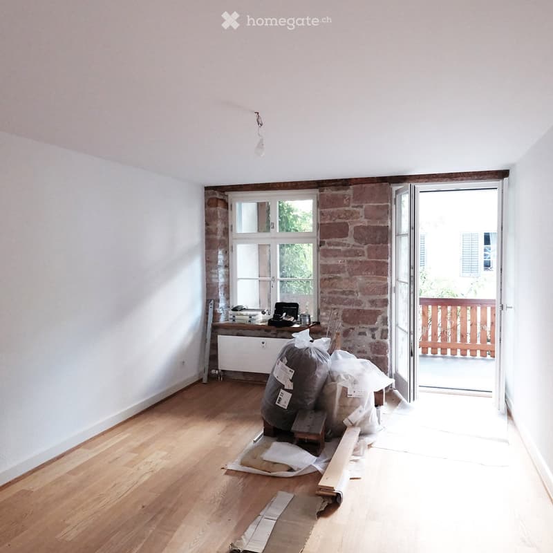 Empty room with wooden flooring, white walls, and a large window. There is a doorway leading to a balcony with wooden railings.