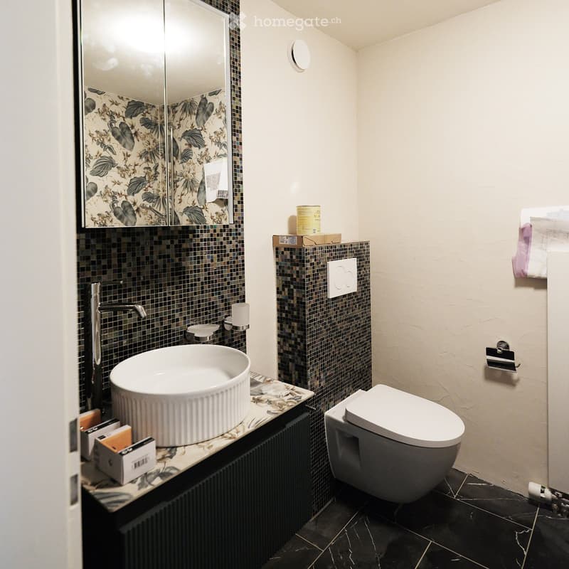 Modern bathroom with a white sink, sleek faucet, floral-patterned mirror, and a contemporary toilet. The walls are tiled with mosaic tiles, and the floor is black marble.