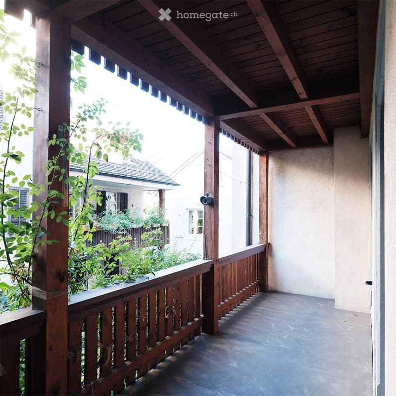 Wooden balcony with a railing, concrete floor, and wooden beams supporting the roof. The balcony overlooks a garden with greenery and a neighboring house.