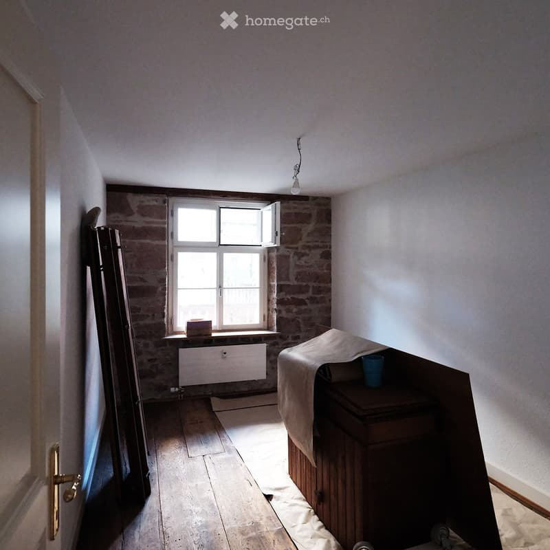 A small, partially furnished room with exposed brick walls, wooden flooring, a window with white frames, a table with a cloth, and a cup on top.