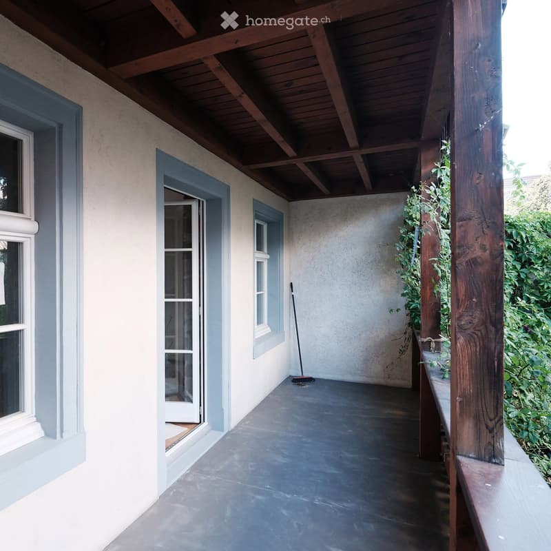 Covered outdoor balcony with wooden ceiling beams, concrete floor, and a small plant in a pot. Features a broom leaning against the wall and a wooden post on the right side.
