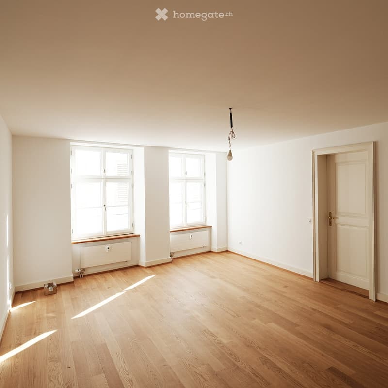 Empty room with light wooden flooring, white walls, two windows with white frames, radiator under each window, and a single hanging light fixture.